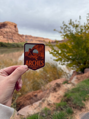 Person holding a 'Arches National Park' sticker with a scenic background of desert landscape.