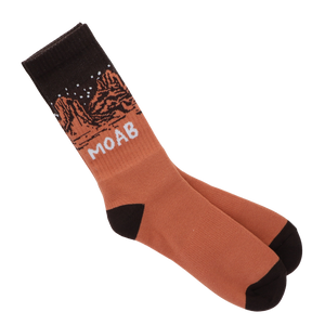 A pair of crew socks in a terracotta rust color with black heels, toes, and cuffs, featuring a printed desert canyon landscape under a starry night sky and the word 'MOAB' in white.