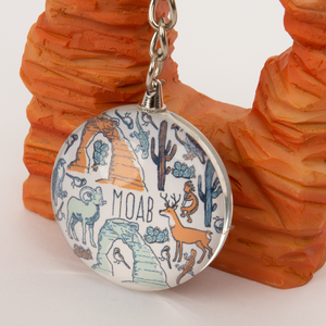 Keychain with desert-themed design featuring animals and 'Moab' text, placed on a wooden surface.