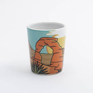 Ceramic mug with a desert scene design featuring a arch and plants on a white background