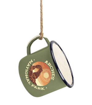 An olive green enamel-style hanging ornament shaped like a mug, suspended by a rope. The front shows a circular logo with a bear and the words 'Nollin' Arches National Park,' and the side has a cream interior with a dark rim.