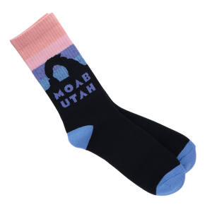 A pair of crew socks on a plain white background. The socks are primarily black with light blue toes and heels, a pink and light pink striped cuff, and a graphic of a mountain silhouette with the text 'MOAB UTAH' in light blue.