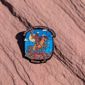 Arches National Park Moab UT enamel pin with Bigfoot riding a rearing unicorn through red rock, lying on red sandstone in Moab, Utah.