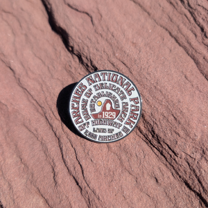 Round white and maroon Arches National Park enamel pin lettered with 1929 and Delicate Arch, lying on red sandstone in Moab, Utah.