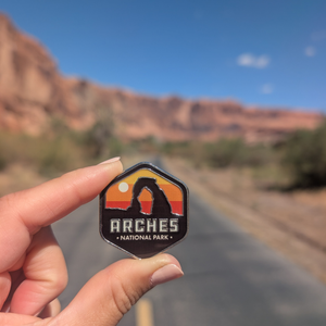 Hand holding the Arches National Park hexagon sunset enamel pin on a road through the Moab red rock cliffs.