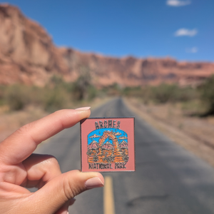 Hand holding the Arches National Park pink sky enamel pin on a road through the Moab red rock cliffs.