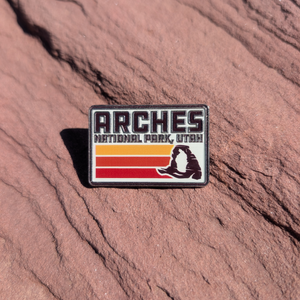 Rectangular Arches National Park Utah enamel pin with retro orange and red stripes and Delicate Arch, lying on red sandstone in Moab, Utah.