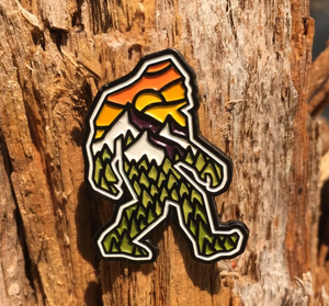Atomic Child enamel pin, a walking Bigfoot filled with green pine trees and an orange mountain sunset, on weathered wood
