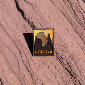 Canyonlands National Park Island in the Sky enamel pin with dark buttes against a golden sky, lying on red sandstone in Moab, Utah.