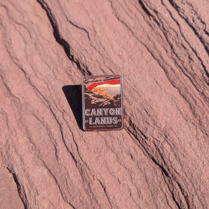 Canyonlands National Park enamel pin framing a sunrise through Mesa Arch, lying on red sandstone in Moab, Utah.