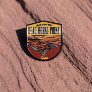 Shield shaped Dead Horse Point enamel pin showing the Colorado River gooseneck below the overlook, lying on red sandstone in Moab, Utah.