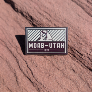 Gray and maroon Moab Utah 1902 enamel pin with Delicate Arch above diagonal stripes, lying on red sandstone in Moab, Utah.