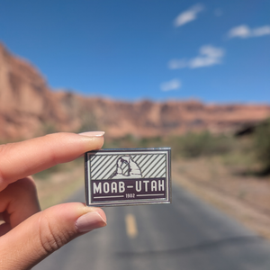 Hand holding the Moab Utah 1902 enamel pin on a road through the Moab red rock cliffs.