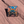 Square Moab enamel pin with a green Jeep climbing red rock under a blue sky, lying on red sandstone in Moab, Utah.