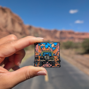 Hand holding the Moab green Jeep enamel pin on a road through the Moab red rock cliffs.