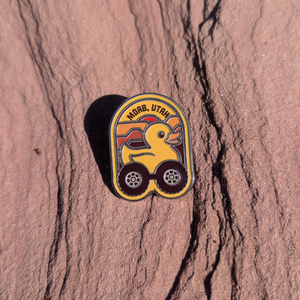 Arch shaped Moab Utah enamel pin with a yellow rubber duck riding on off road tires, lying on red sandstone in Moab, Utah.