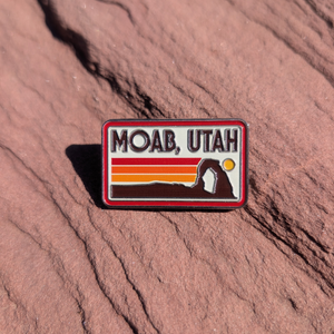 Rectangular Moab Utah enamel pin with retro sunset stripes and a Delicate Arch silhouette, lying on red sandstone in Moab, Utah.