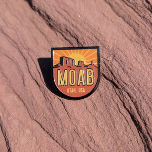 Shield shaped Moab Utah USA enamel pin with a sunburst rising behind red mesas, lying on red sandstone in Moab, Utah.