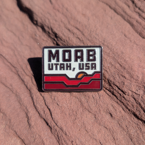 Rectangular Moab Utah USA enamel pin with red rock waves and a setting sun, lying on red sandstone in Moab, Utah.