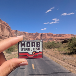 Hand holding the Moab Utah USA red rock enamel pin on a road through the Moab red rock cliffs.