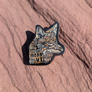 Die cut enamel pin of a wolf head in detailed line art with Moab UT lettering, lying on red sandstone in Moab, Utah.