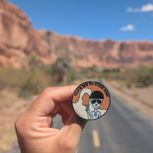 Hand holding the Take Me To Your Dirt alien enamel pin on a road through the Moab red rock cliffs.